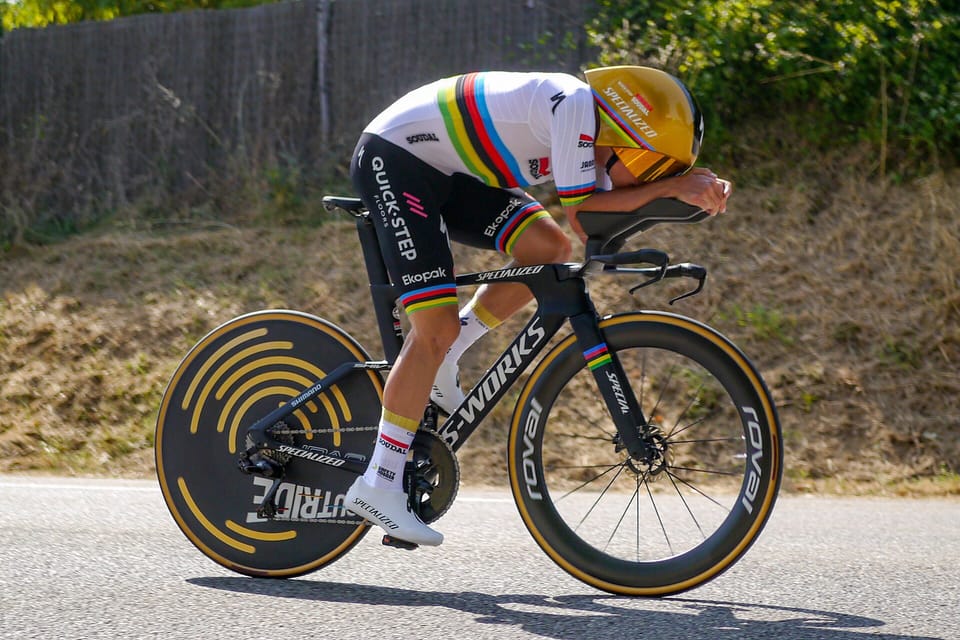 A rider in a rainbow-striped jersey and gold helmet on a time-trial bike with a disc wheel