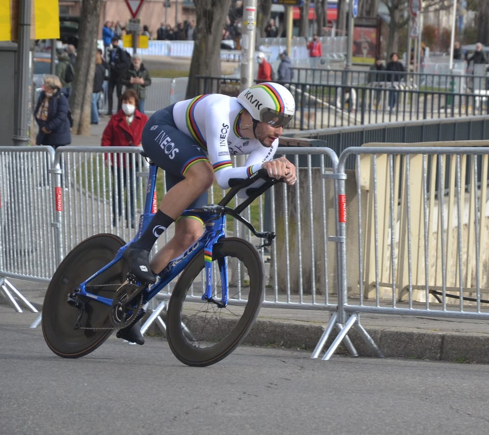 Filippo Ganna in a white rainbow-banded world champion’s skinsuit and aero helmet, riding a time-trial bike past crowd barriers