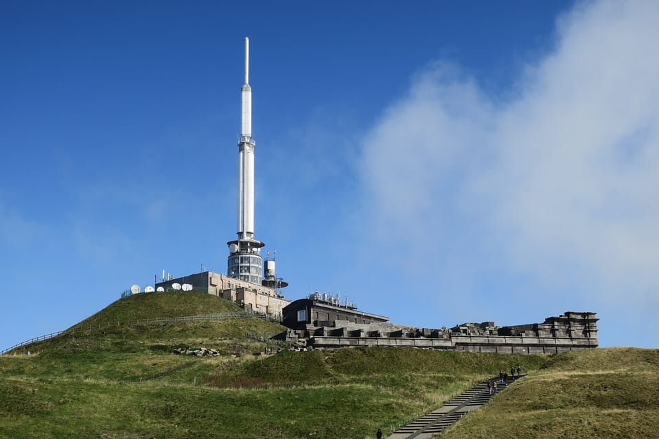 The grassy summit of the Puy de Dôme with a tall white transmitter mast, buildings and the stone ruins of a Roman temple under a blue sky