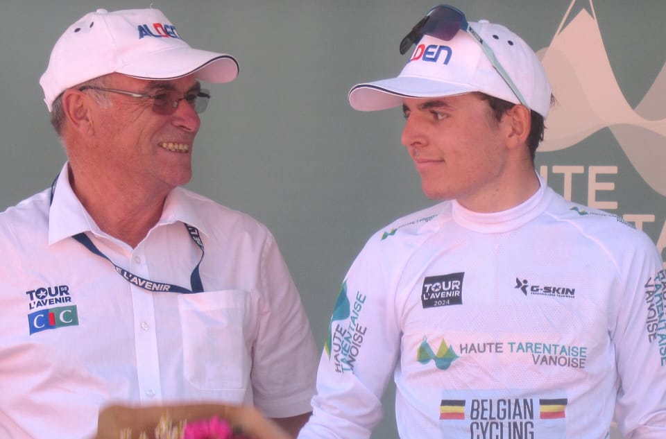 A young rider in a white jersey and cap smiles on a podium beside an older man in a white cap