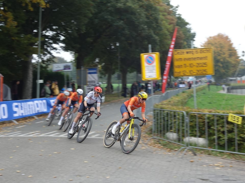 A group of riders on a road section of a gravel race, passing barriers and spectators