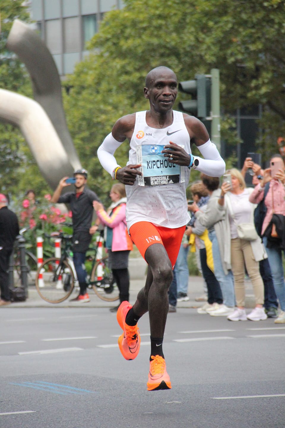 Eliud Kipchoge running in a white vest and orange shoes during the 2022 Berlin Marathon, with spectators behind barriers