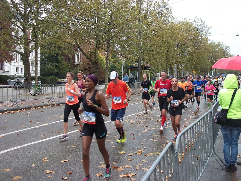 Runners on a tree-lined street scattered with autumn leaves, with spectators behind a barrier on the right