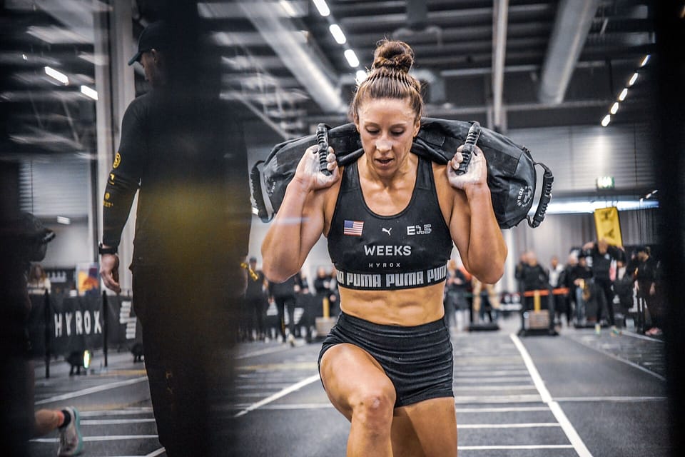 A female athlete in a black crop top and shorts lunges along an indoor race lane with a sandbag across her shoulders