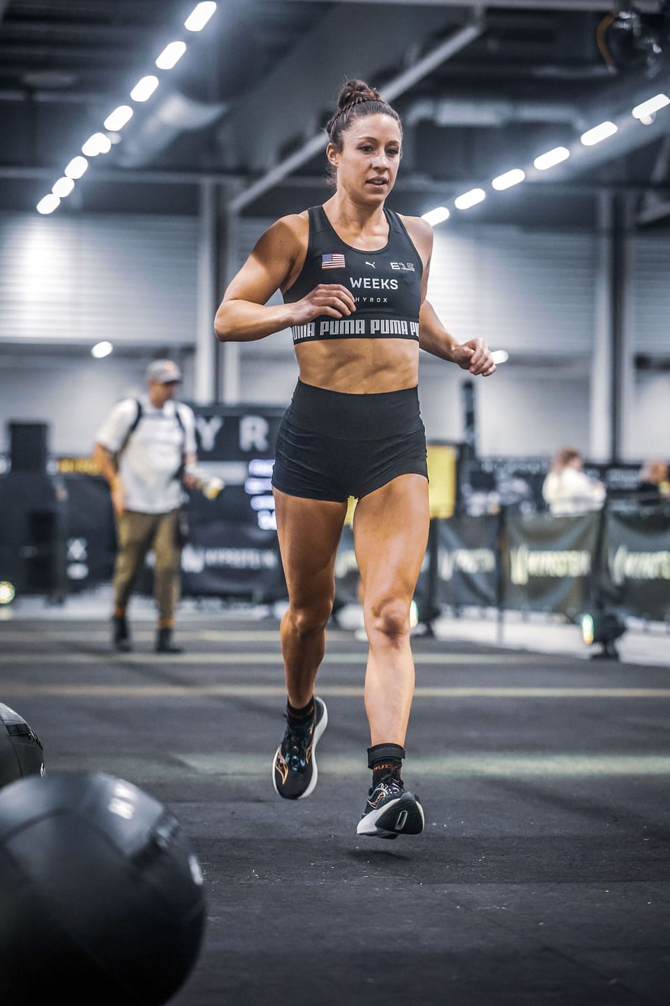 A female athlete in black running kit jogs across an indoor arena floor past a medicine ball