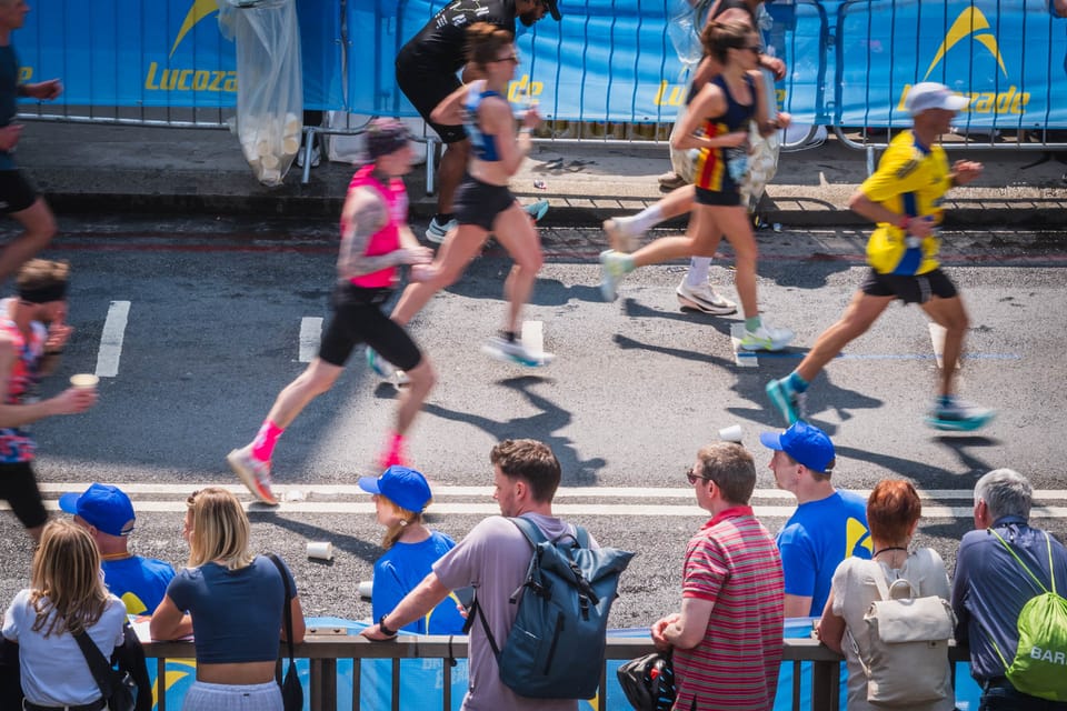 Runners in colourful kit pass a drinks station while spectators lean on a railing in the foreground