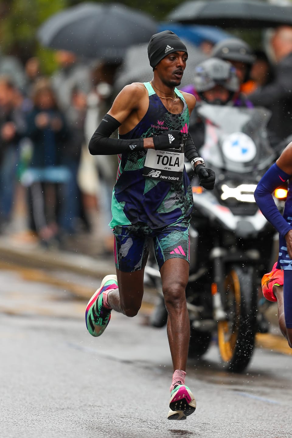 A runner in a dark vest and beanie with a TOLA race bib running on a wet road in the rain, with a motorbike behind him