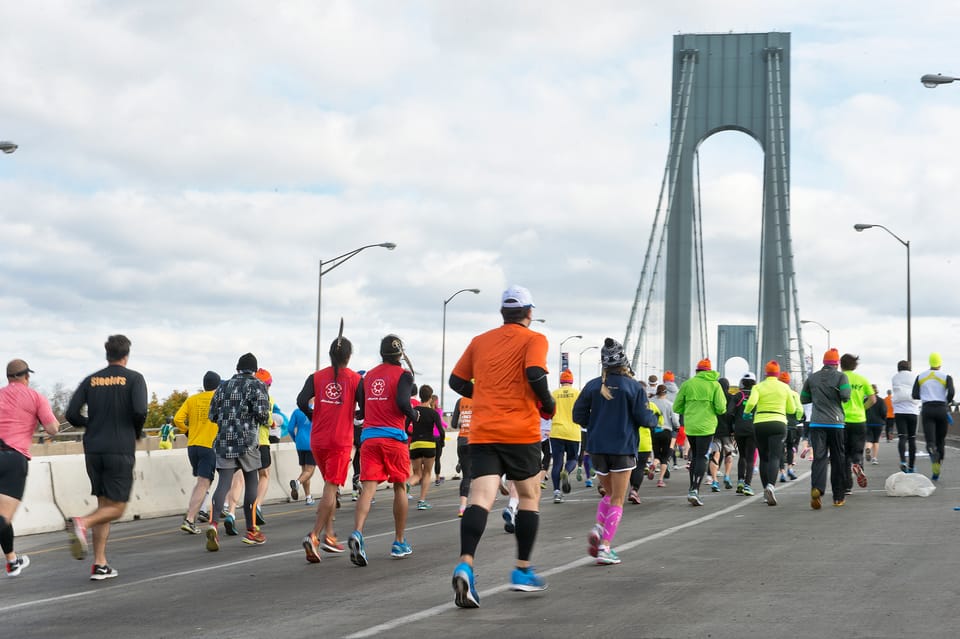 Marathon runners crossing a wide road with the towers of the Verrazzano-Narrows Bridge behind them