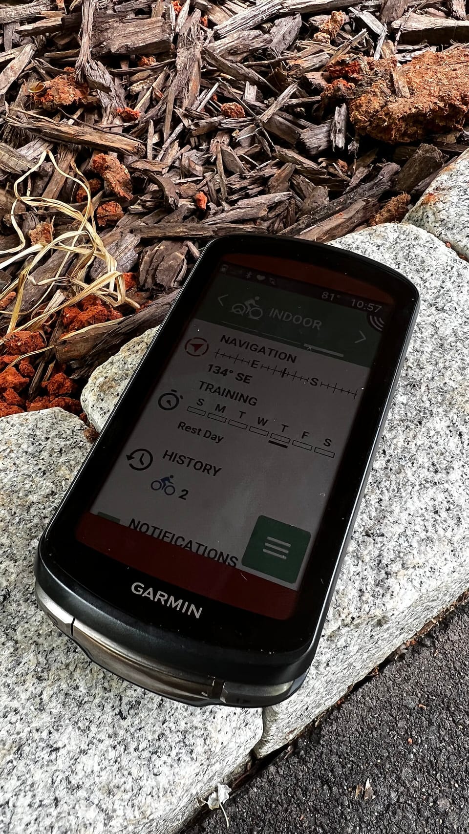 A black Garmin bike computer with a touchscreen showing navigation and training widgets, resting on a granite kerb
