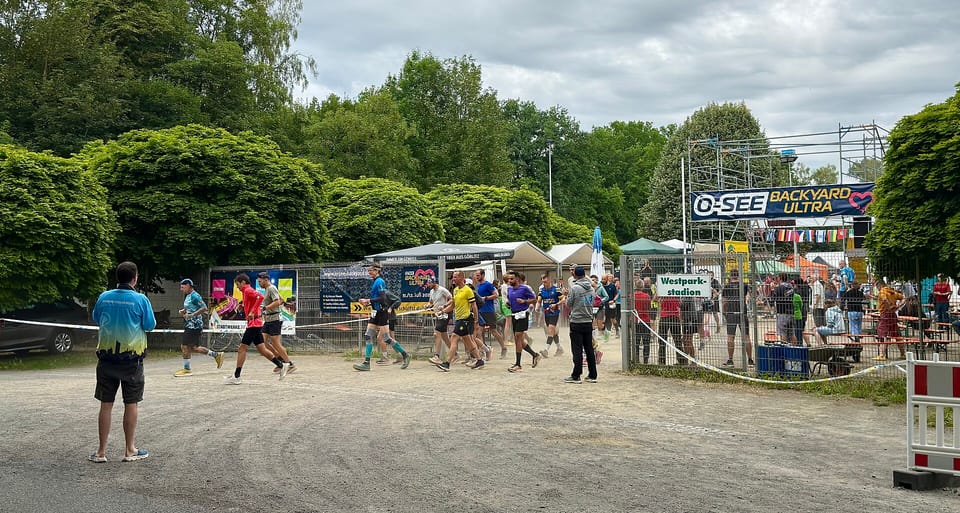 Runners leave a fenced start area under an O-SEE Backyard Ultra banner, with trees behind