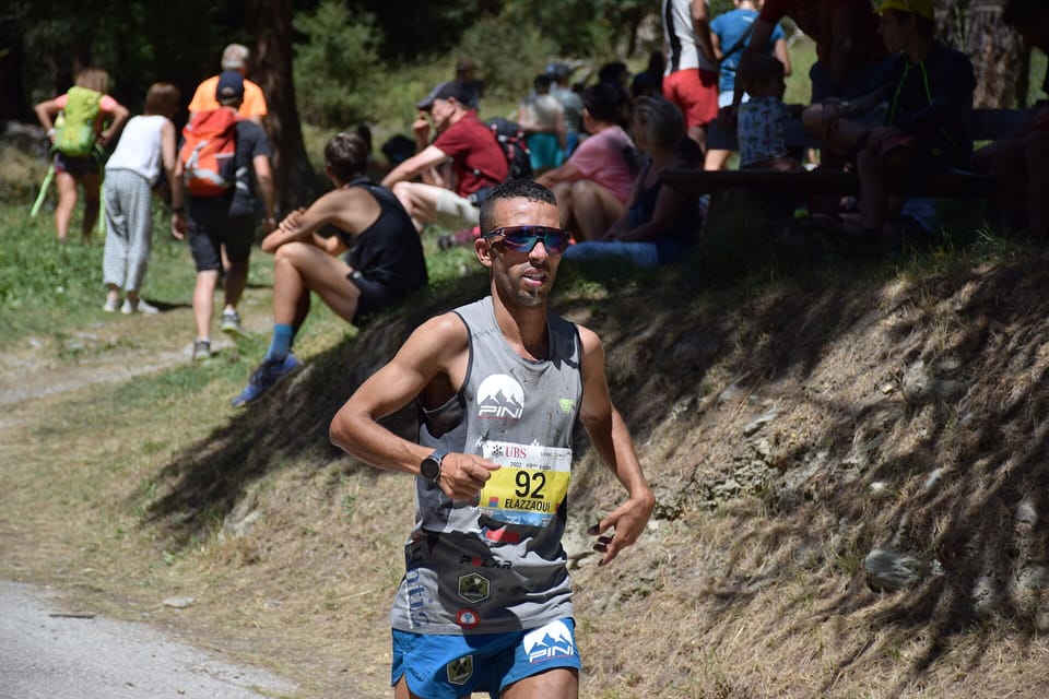 A runner in sunglasses and race number 92 runs up a dirt path past spectators sitting on a grassy bank