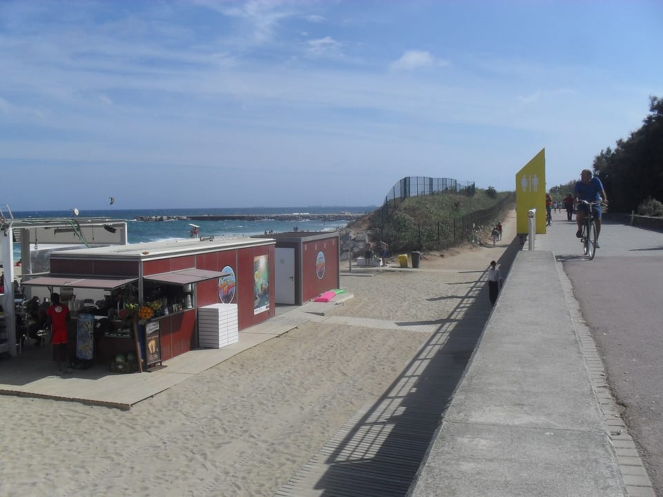 A cyclist on a seafront promenade beside a sandy beach with a kiosk and the sea beyond