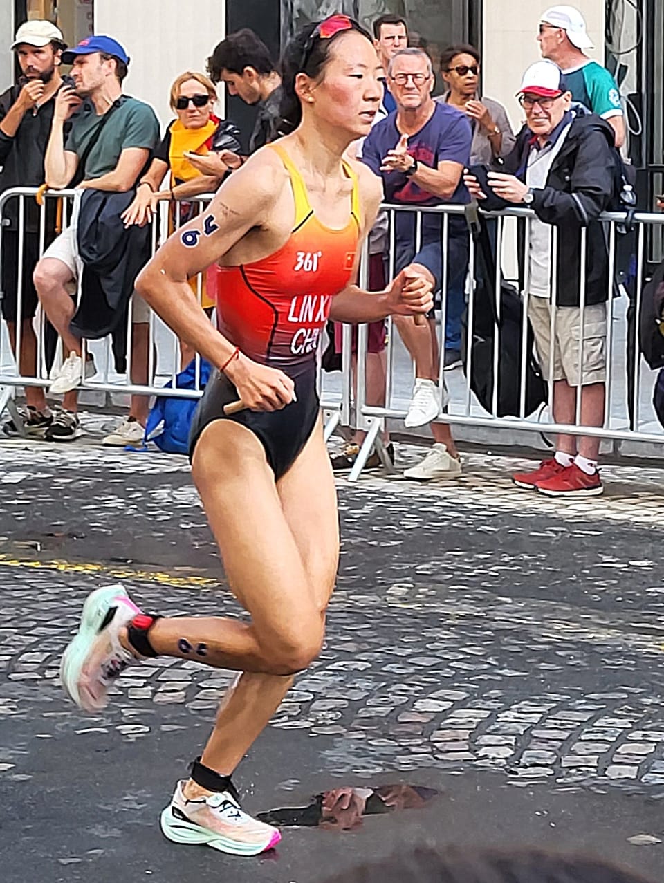 Xinyu Lin in an orange and black China trisuit running on a cobbled street past spectators behind barriers