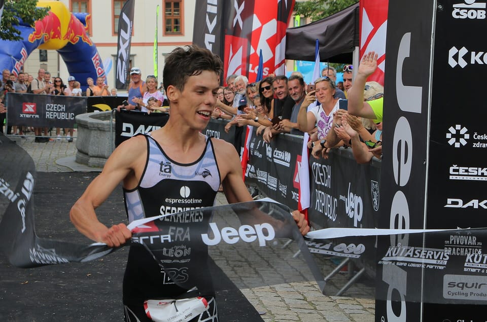 A triathlete in a black race suit takes the finishing tape between crowd barriers at an Xterra race
