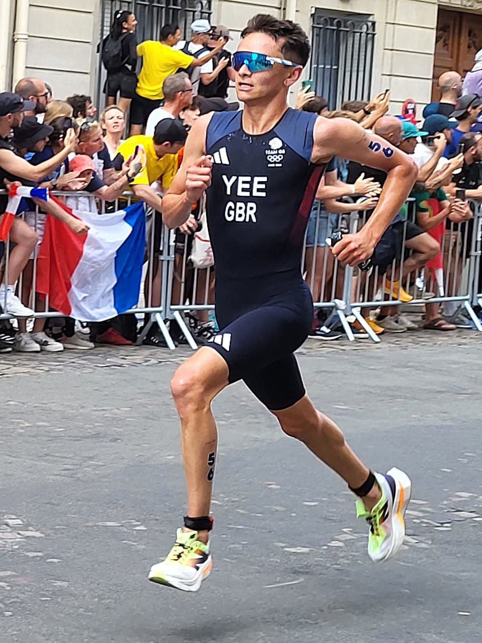 Alex Yee in a navy Great Britain trisuit and blue sunglasses running past a crowd behind barriers