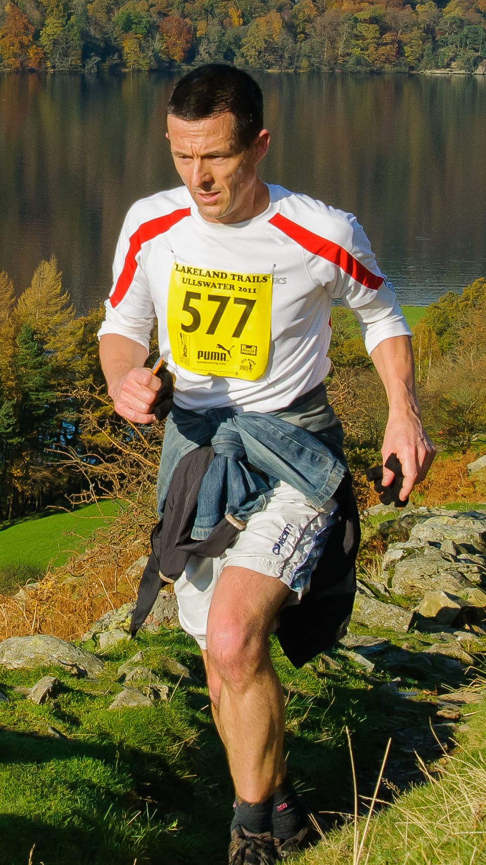 A trail runner in a race number running on a grassy hillside above a lake