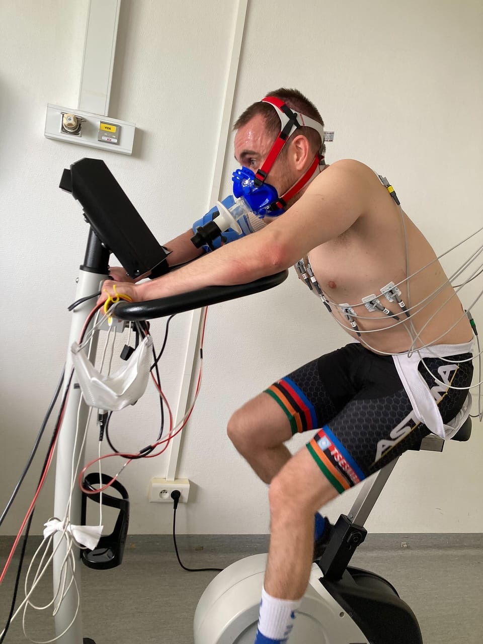 A cyclist on an indoor bike wearing a breathing mask connected to gas-analysis equipment during a laboratory fitness test